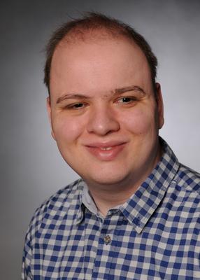 The portrait shows a man with short, brown hair, which is already thinning slightly at the top of his head. He has a round face with a high forehead, well-defined dark eyebrows, brown eyes and light-coloured skin. He has a friendly expression with a slight smile. He wears a blue and white chequered shirt. This image description was partly created with the help of artificial intelligence.